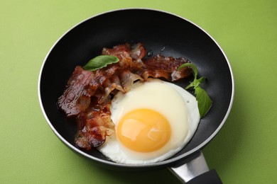 Fried egg, bacon and basil in frying pan on green background