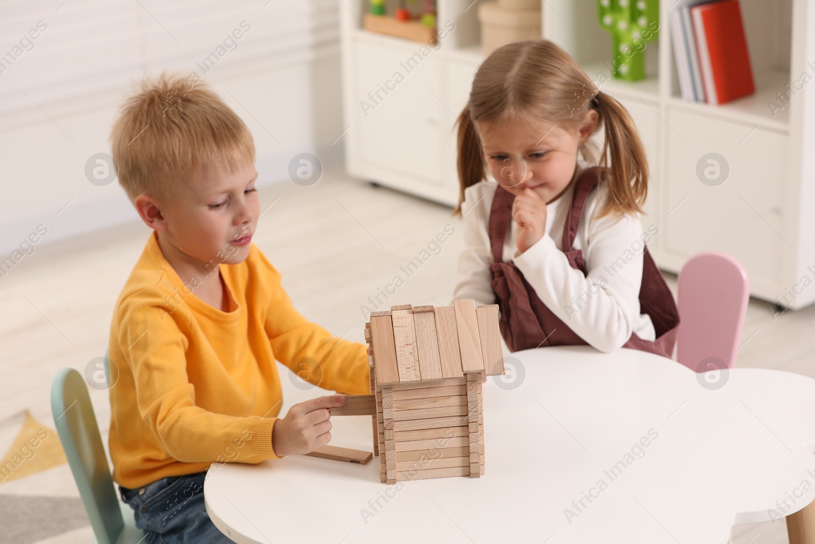 Photo of Little boy and girl playing with wooden house at white table indoors. Children's toys