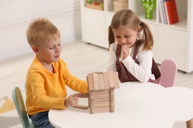 Little boy and girl playing with wooden house at white table indoors. Children's toys