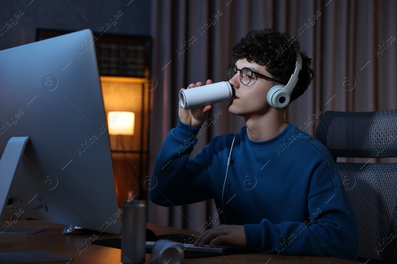 Photo of Young man with energy drink and headphones playing video game at wooden desk indoors