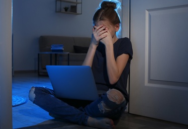 Photo of Frightened teenage girl with laptop on floor in dark room. Danger of internet