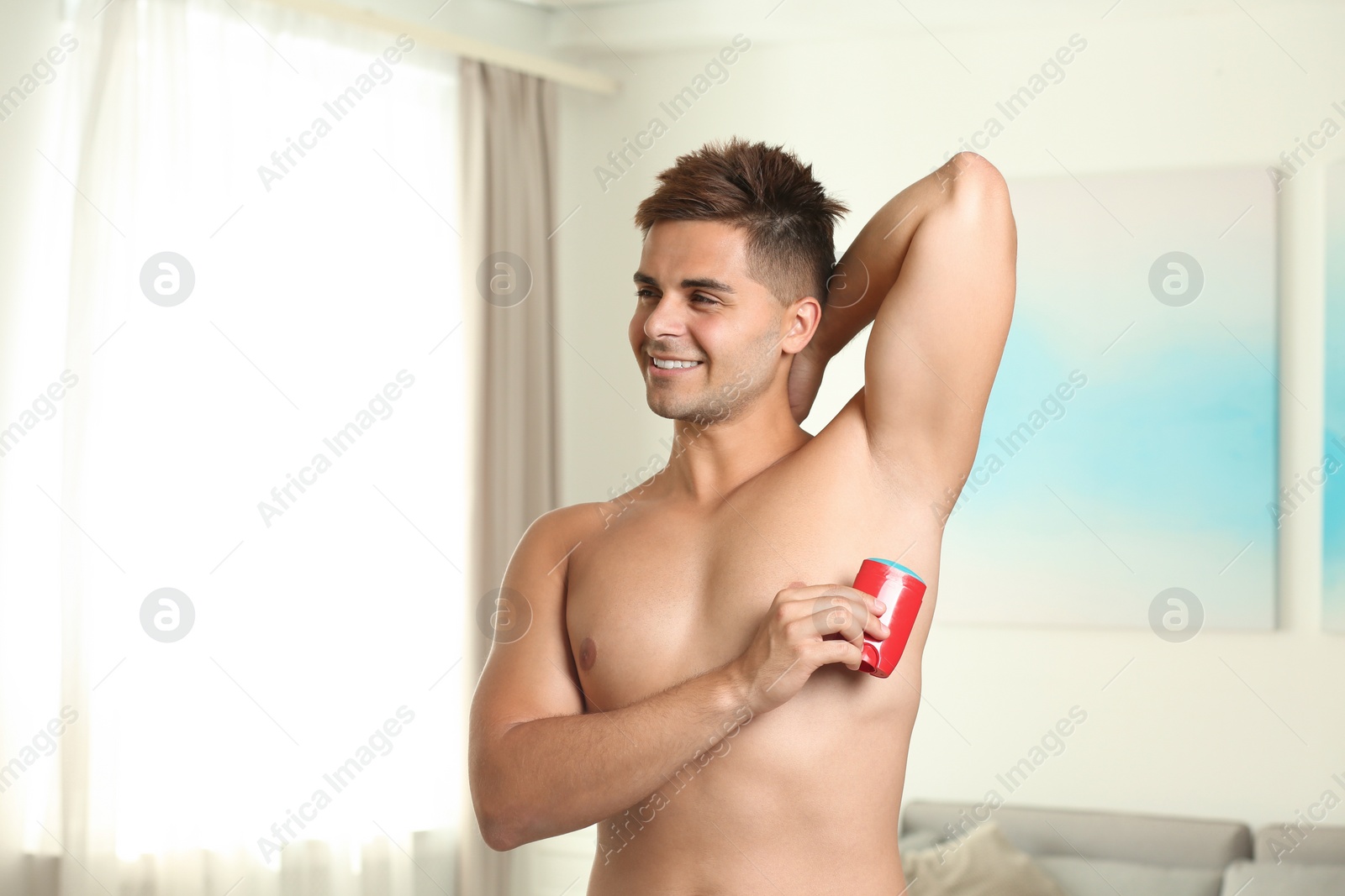 Photo of Young man applying fresh deodorant to armpit indoors