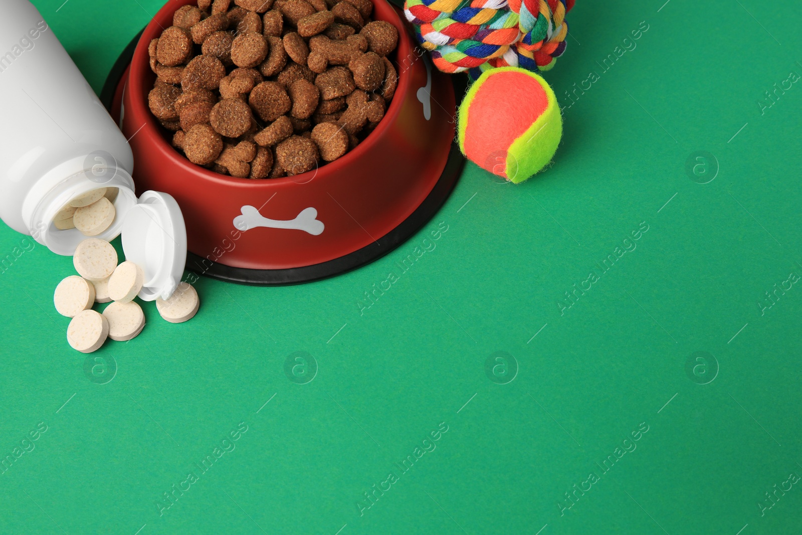 Photo of Bowl with dry pet food, bottle of vitamins and toys on green background. Space for text