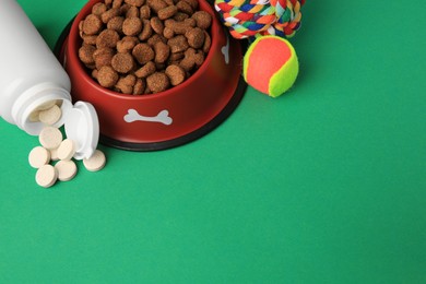 Bowl with dry pet food, bottle of vitamins and toys on green background. Space for text