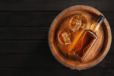 Whiskey with ice cubes in glasses, bottle and barrel on black wooden table, top view. Space for text