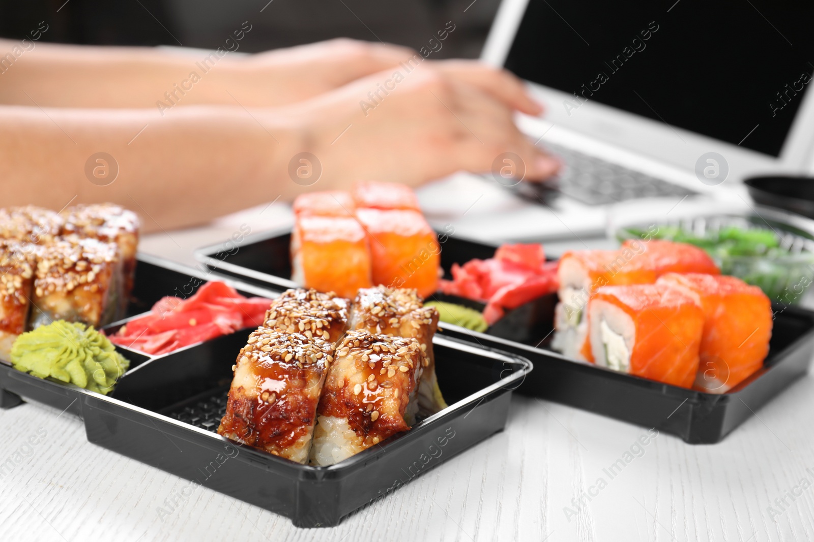 Photo of Boxes with different sushi rolls and blurred woman using laptop on background. Food delivery