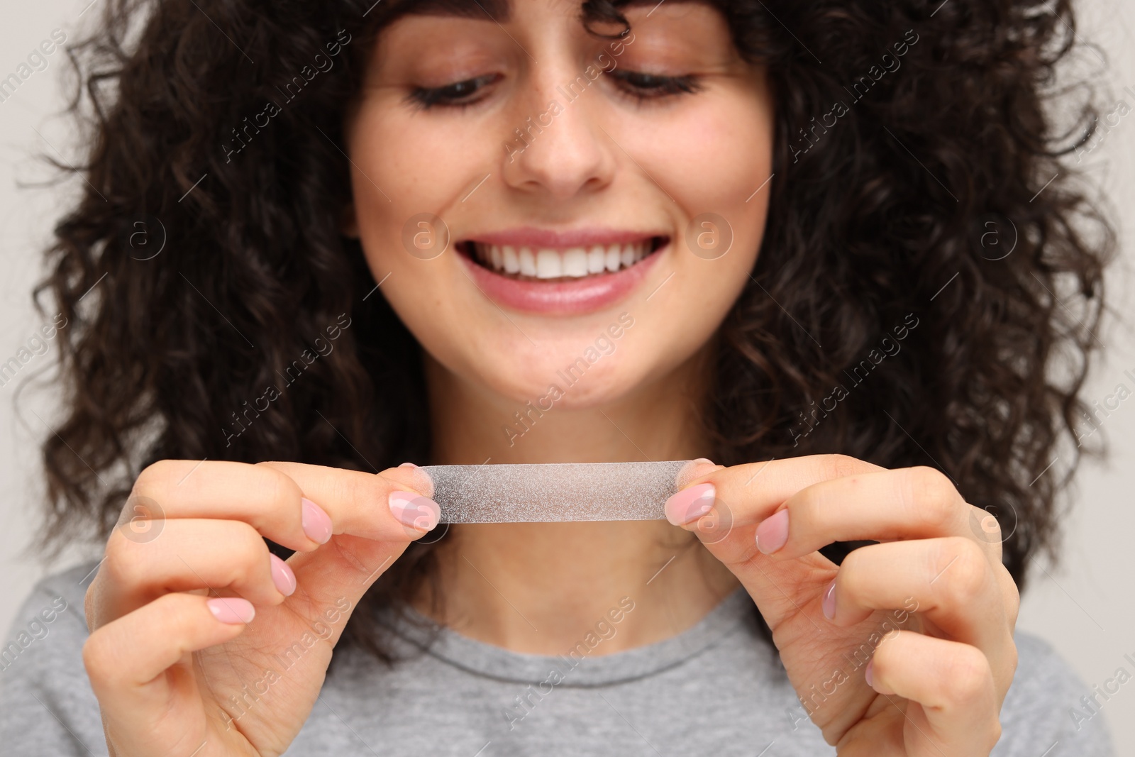 Photo of Young woman holding teeth whitening strip on light grey background, closeup