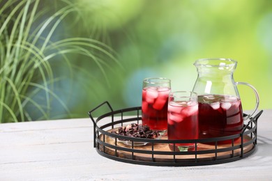 Photo of Refreshing hibiscus tea with ice cubes and dry roselle flowers on white wooden table against blurred green background. Space for text