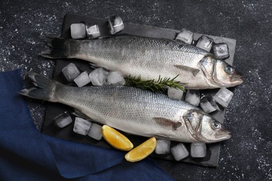 Fresh raw sea bass fish, ice cubes and lemon wedges on black table, flat lay