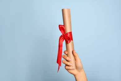 Photo of Student holding rolled diploma with red ribbon on light blue background, closeup