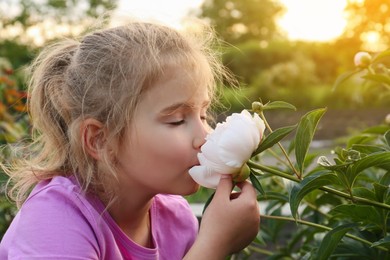 Photo of Cute little girl sniffing blooming peony outdoors