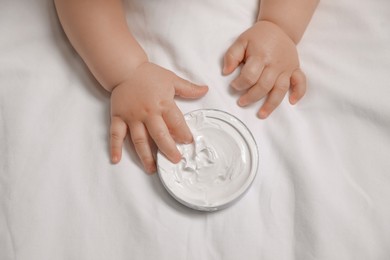 Photo of Cute little baby playing with jar of moisturizing cream on bed, closeup