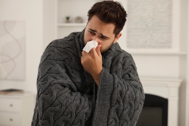 Photo of Sick man wrapped in blanket with tissue blowing nose at home. Cold symptoms