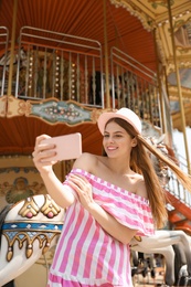 Photo of Young pretty woman taking selfie near carousel in amusement park