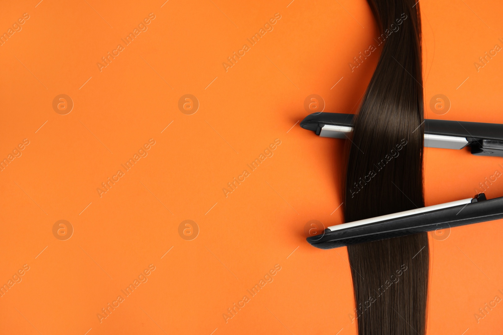 Photo of Straightener with brown hair lock on orange background, flat lay. Space for text