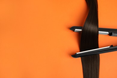 Photo of Straightener with brown hair lock on orange background, flat lay. Space for text