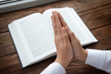 Woman holding hands clasped while praying at wooden table with Bible, closeup