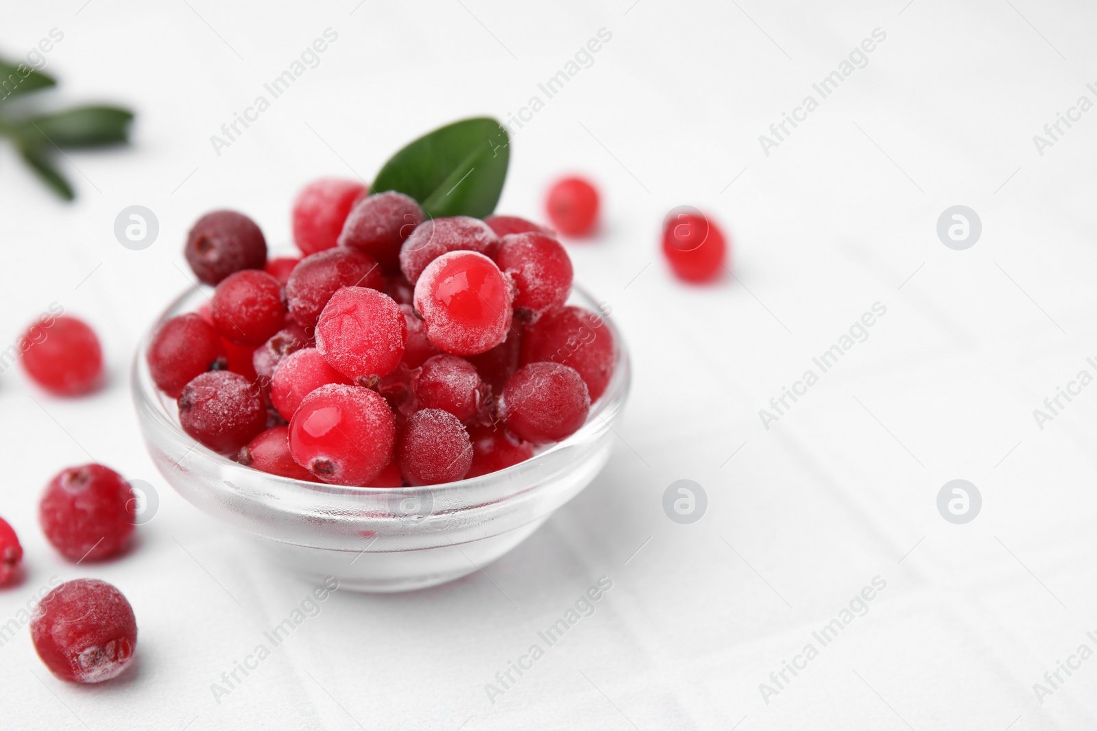 Photo of Frozen red cranberries in bowl and green leaves on white tiled table, closeup. Space for text