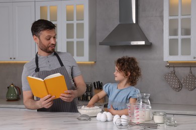 Photo of Cute little girl and her father with recipe book cooking in kitchen