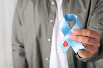 Photo of Man holding light blue ribbon with paper blood drop indoors, closeup. World Diabetes Day