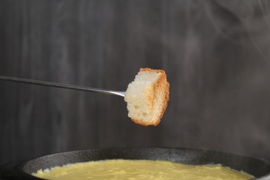 Photo of Dipping piece of bread into fondue pot with tasty melted cheese against dark gray background, closeup