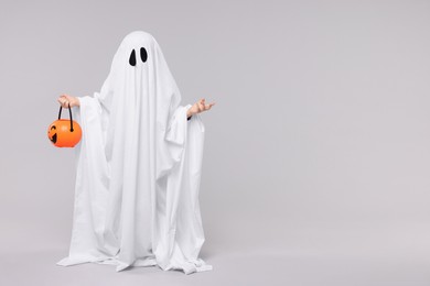 Child in white ghost costume holding pumpkin bucket on light grey background, space for text. Halloween celebration