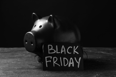 Photo of Piggy bank and card with words Black Friday on table against dark background