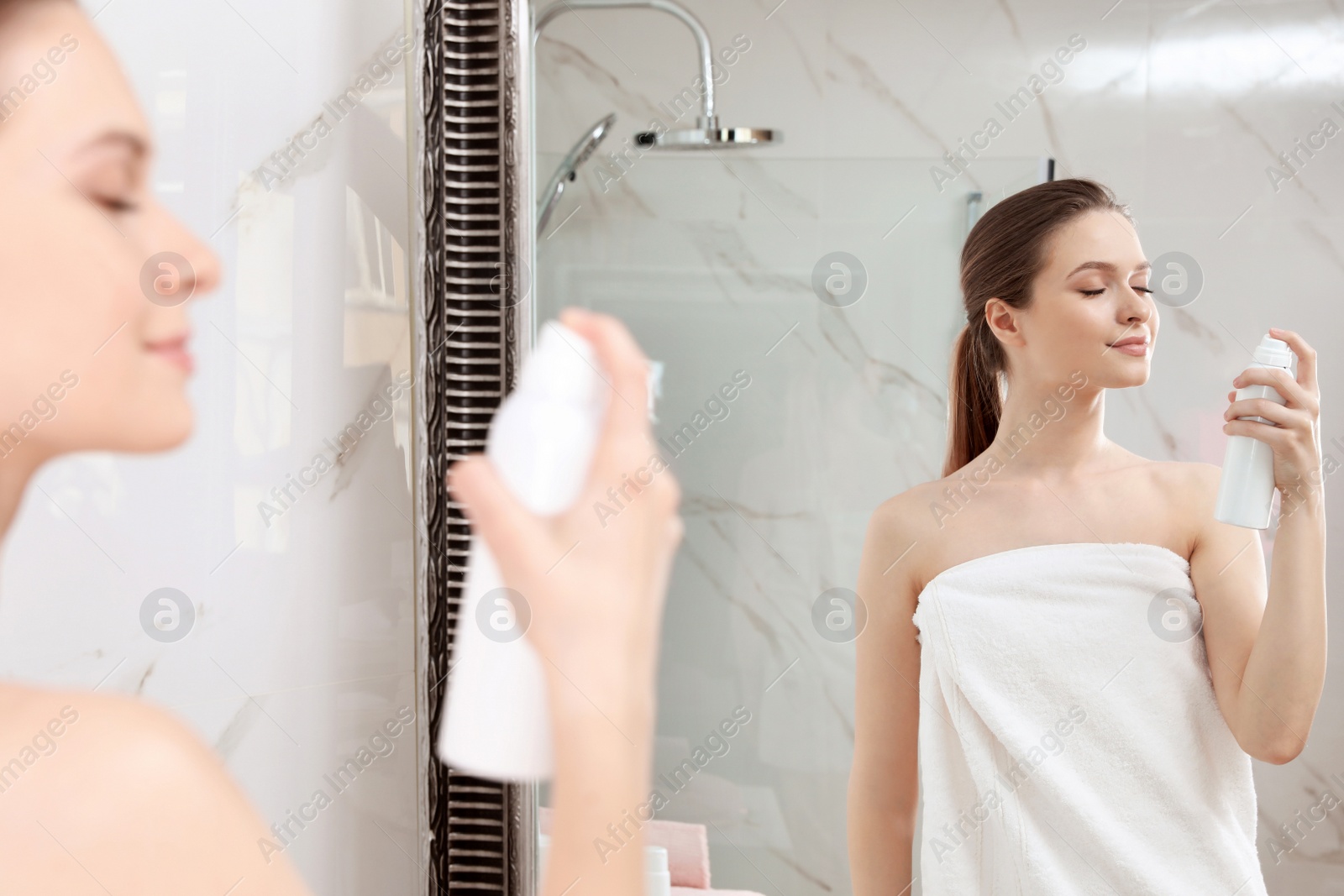 Photo of Young woman applying thermal water on face near mirror in bathroom. Cosmetic product