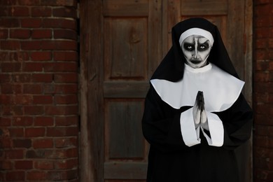 Photo of Portrait of scary devilish nun with clasped hands near red brick wall, space for text. Halloween party look