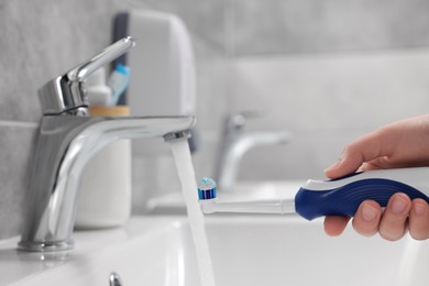 Photo of Woman holding electric toothbrush with paste near flowing water above sink in bathroom, closeup