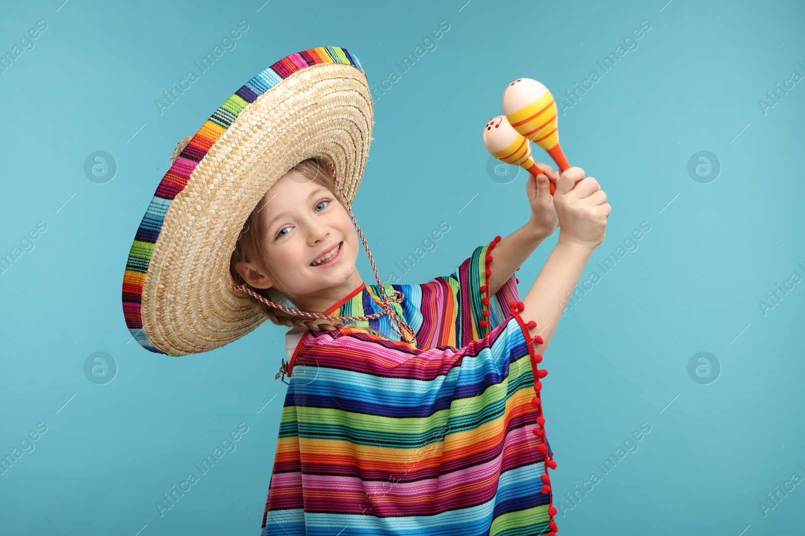 Photo of Cute girl in Mexican sombrero hat and poncho dancing with maracas on light blue background