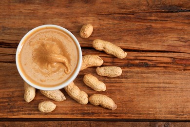 Photo of Delicious nut butter in bowl and peanuts on wooden table, top view