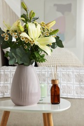 Aromatic reed air freshener and vase with bouquet on white table in bedroom