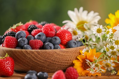 Photo of Bowl with different fresh ripe berries and beautiful flowers on table outdoors, closeup