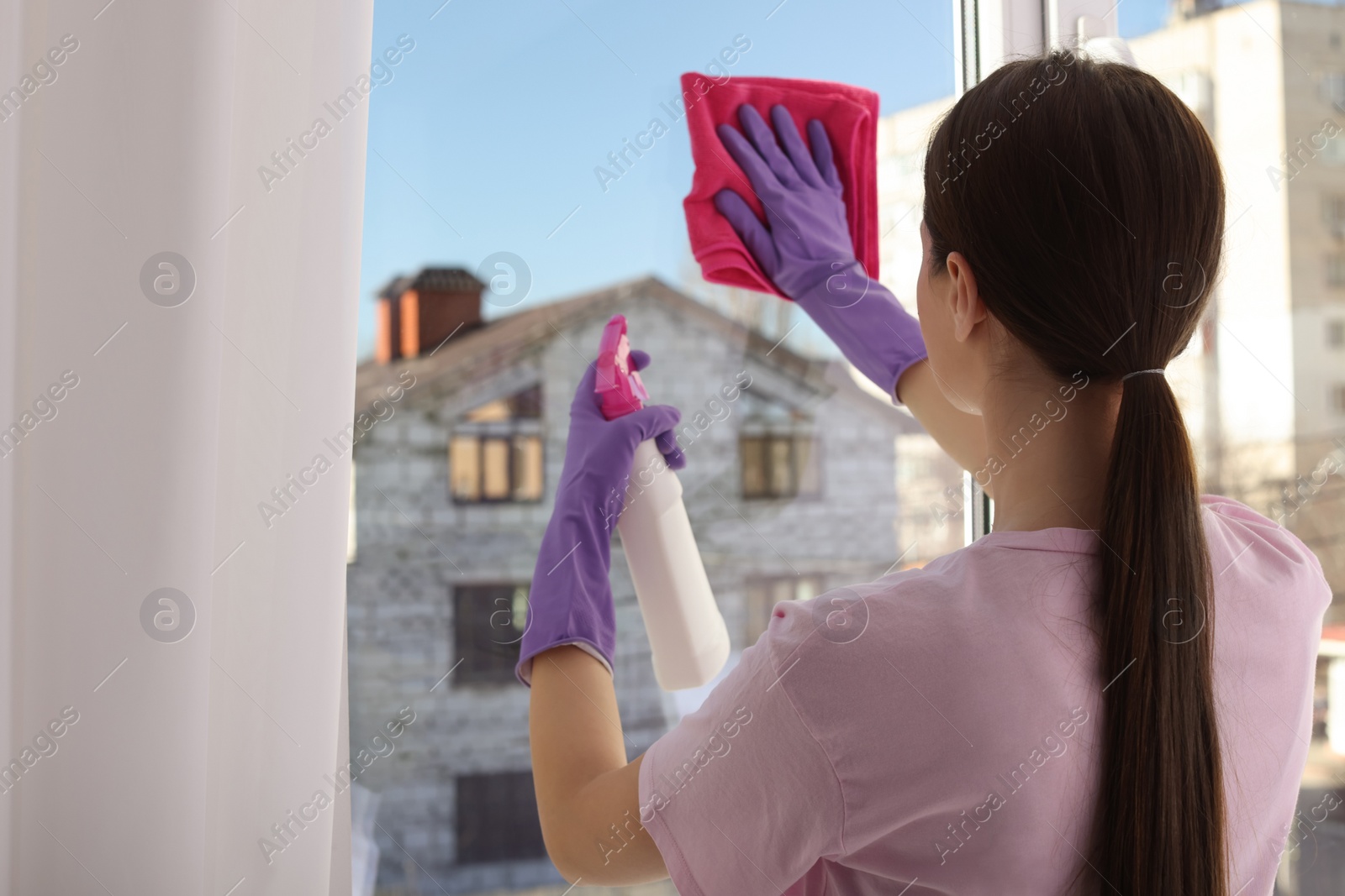 Photo of Young woman cleaning window glass with rag and detergent at home, back view. Space for text