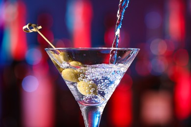 Pouring cocktail into martini glass with olives on blurred background, closeup