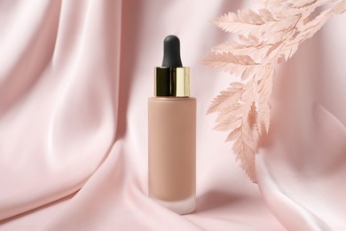 Photo of Bottle of skin foundation and decorative plant on pink cloth. Makeup product