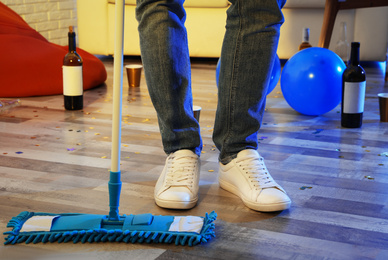 Photo of Young man with mop cleaning messy room after party, closeup of legs