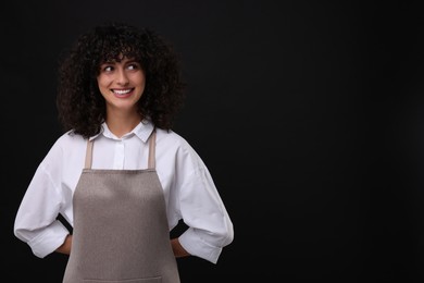 Photo of Happy woman wearing kitchen apron on black background, space for text. Mockup for design