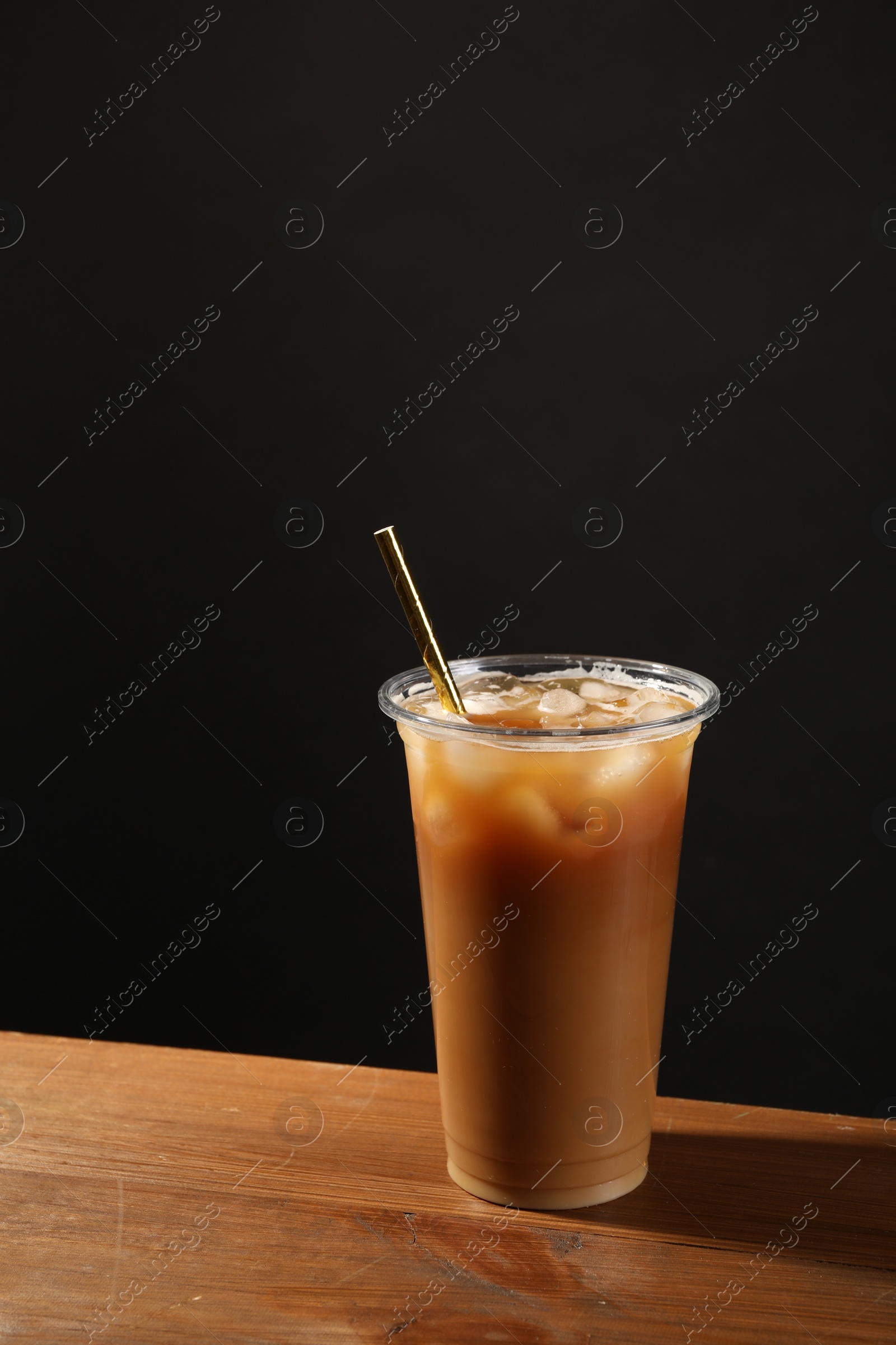 Photo of Refreshing iced coffee with milk in takeaway cup on wooden table against black background, space for text