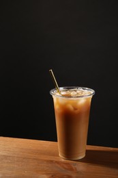 Photo of Refreshing iced coffee with milk in takeaway cup on wooden table against black background, space for text