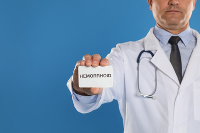 Doctor holding business card with word HEMORRHOID on blue background, closeup