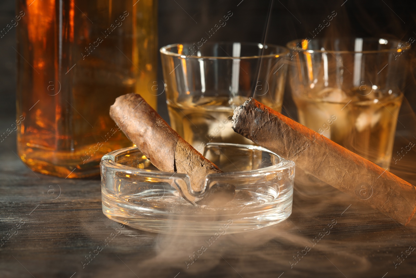 Photo of Cigars, ashtray and whiskey with ice cubes on black wooden table, closeup