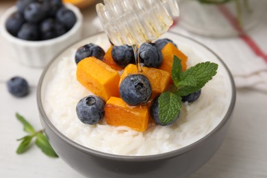 Photo of Pouring honey onto delicious rice porridge with blueberries, pumpkin and mint on table, closeup