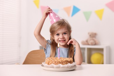 Cute girl in party hat with birthday cake at table indoors