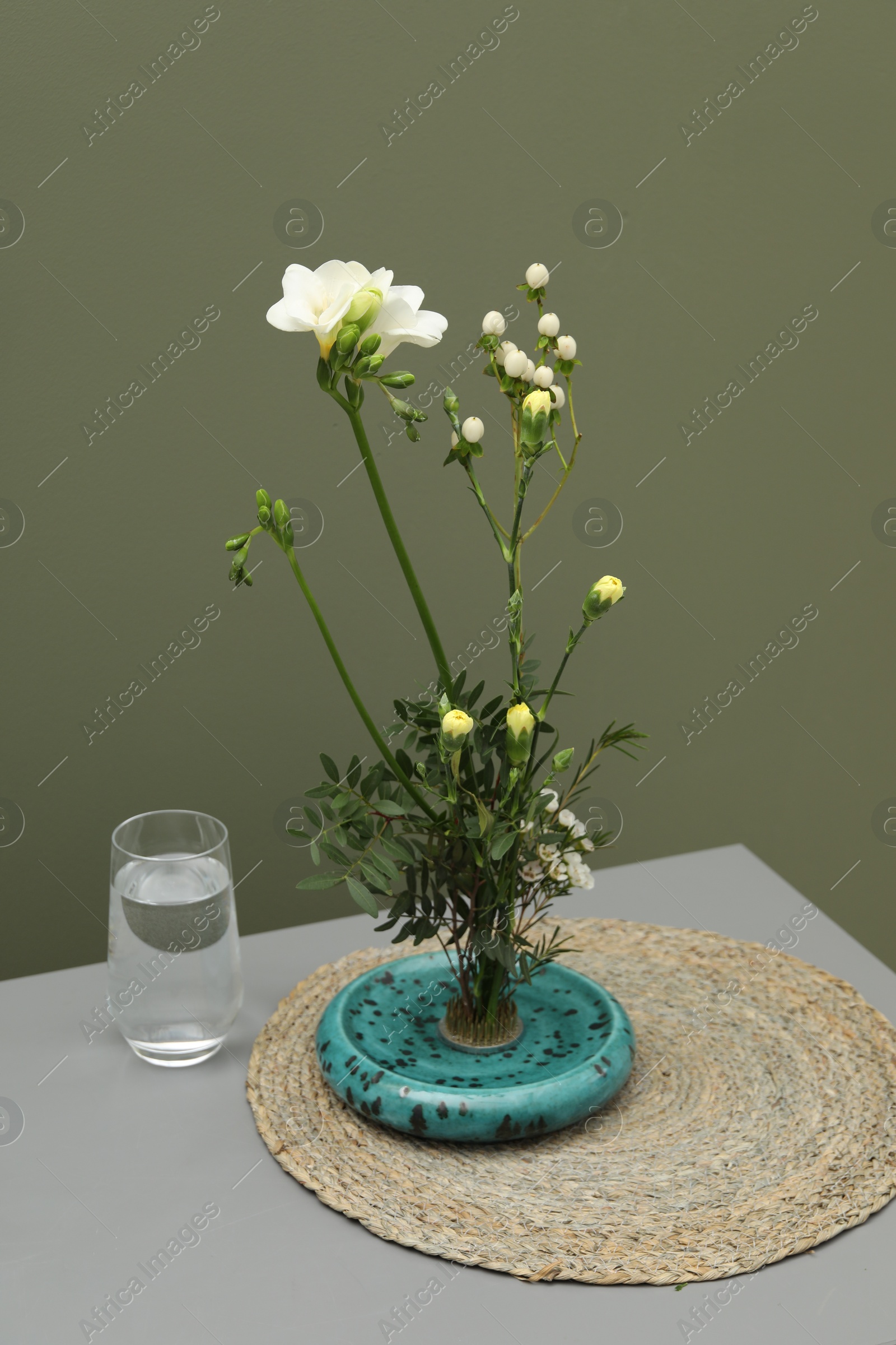 Photo of Stylish ikebana with beautiful flowers, green branches and glass of water on gray table near olive wall