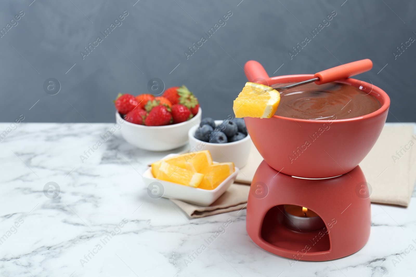 Photo of Fondue pot with melted chocolate, fresh orange, different berries and fork on white marble table. Space for text