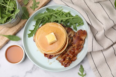 Tasty pancakes with butter, fried bacon and fresh arugula on white marble table, flat lay