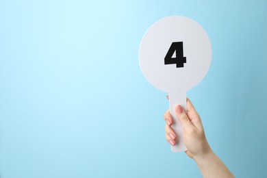 Photo of Woman holding auction paddle with number 4 on light blue background, closeup. Space for text