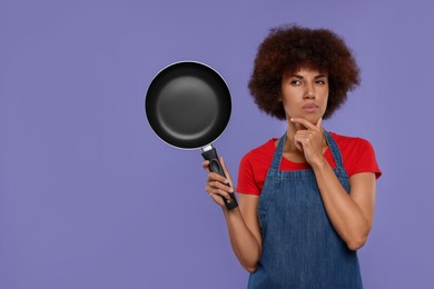 Photo of Thoughtful young woman in apron holding frying pan on purple background. Space for text